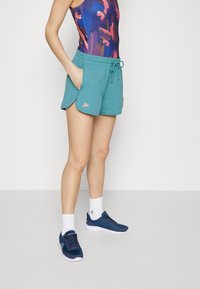 Un short turquoise avec une taille à cordon, comportant des poches latérales et un logo rose. Porté avec un haut sans manches coloré et des chaussures de sport bleues.