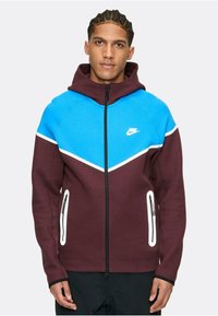 Sudadera Nike en burdeos y azul. Presenta bloques de color en ángulo, cremallera frontal, capucha y bolsillos con cremallera blanca. Fabricada con tejido suave.