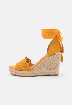 Sandalia cuña de ante amarillo con diseño de recortes florales, suela de yute trenzado y lazos de cinta en el tobillo. Presenta una punta redondeada y tacón texturizado.