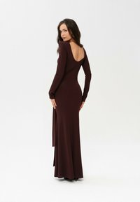 Długa burgundowa sukienka uszyta z gładkiego materiału, z długimi rękawami, głębokim dekoltem na plecach oraz bocznym rozcięciem.