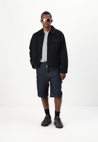 Svart jakke med krage, grå t-skjorte, denimshorts og joggesko. Tilbehøret inkluderer rektangulære sølvfargede solbriller.