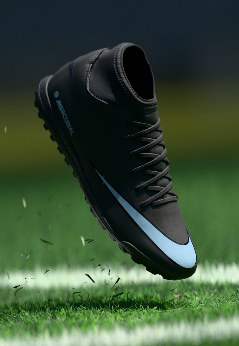 Botín de fútbol negro con diseño de caña alta, que presenta una franja de acento azul claro, superficie texturizada y cierre con cordones, colocado sobre césped.