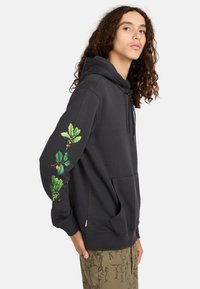 Jongere met lange, krullende haren draagt een zwarte hoodie met groene botanische bladeren en fruitafdrukken op de rechtermouw.