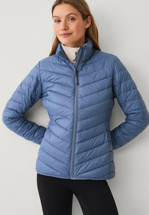 Vrouw draagt een lichtblauwe doorgestikte jas over een beige top met rits, staand met handen achter de rug tegen een effen achtergrond.