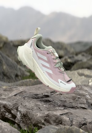 Scarpa da trekking rosa chiaro e bianca con suola spessa e sistema di allacciatura rapida che fluttua sopra un terreno roccioso con montagne sfocate sullo sfondo.