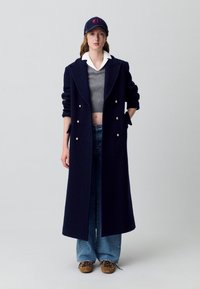 Cappotto lungo in lana navy con bottoni dorati, abbinato a un maglione grigio corto, jeans blu e un cappello navy con logo rosso.