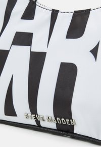 Μαύρη και λευκή τσάντα υφάσματος με έντονο γραφικό κείμενο. Υφή με υφασμάτινη επιφάνεια και ασημένιο λογότυπο "Steve Madden" ενσωματωμένο στο μπροστινό μέρος.