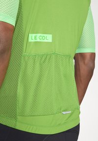 Zeleni biciklistički dres s mrežastim dijelovima, teksturiranom tkaninom i logotipom "LE COL". Ima kratke rukave i stražnji džep.