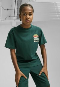 Camiseta de algodón verde oscuro de manga corta, con un logo gráfico multicolor "Puma Board Rebels" en el pecho.