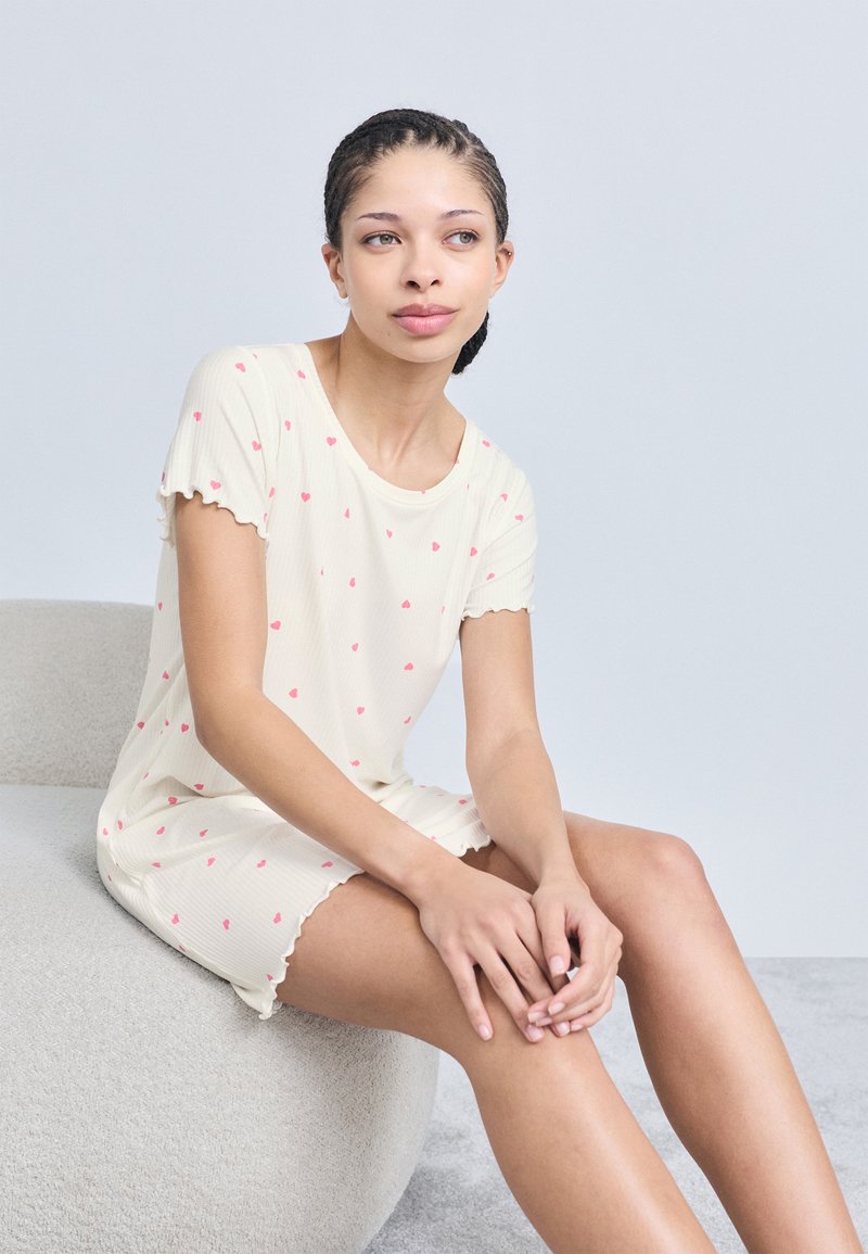 Φόρεμα κοντό μανίκι σε ανοιχτό κίτρινο χρώμα με μικρές ροζ καρδιές, ριμπ ύφανση, κυματιστά περιθώρια, μήκος ως το γόνατο, εφαρμοστή γραμμή, φορεμένο ενώ κάθεται.