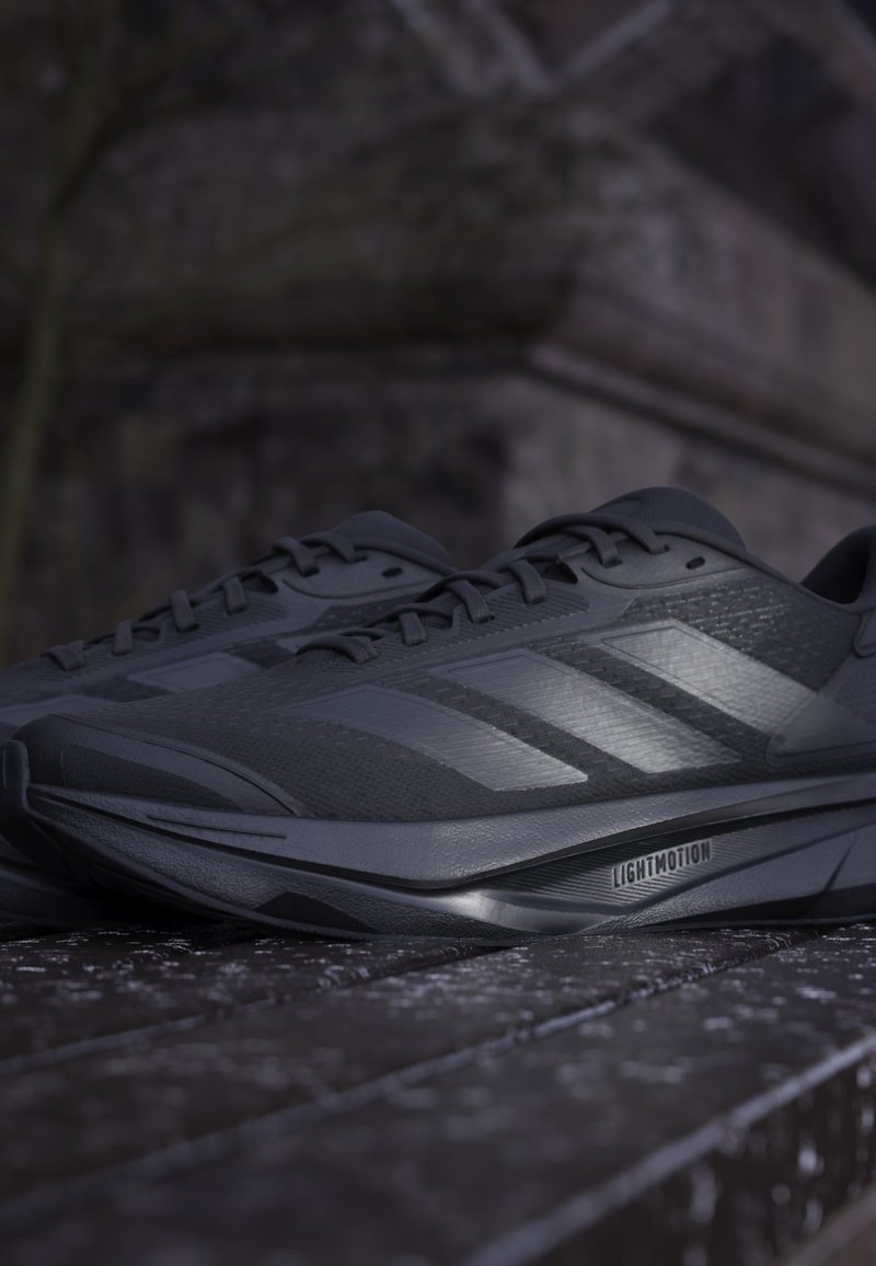 Paire de chaussures de course Adidas noires avec semelles Lightmotion reposant sur une surface sombre texturée en faible lumière.
