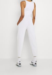 Person klädd i en vit ärmlös topp och högmidjade vita joggers med fickor, står med händerna i fickorna, vänd bort.