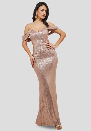 Vrouw die een nauwsluitende, off-shoulder galajurk van roségouden pailletten draagt, met geometrische oorbellen en open-toe hakken, poserend tegen een effen achtergrond.