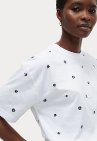 Witte katoenen t-shirt met verspreide zwart bloemenborduursels. Korte mouwen en een ronde halslijn. Zachte textuur, ontspannen pasvorm.