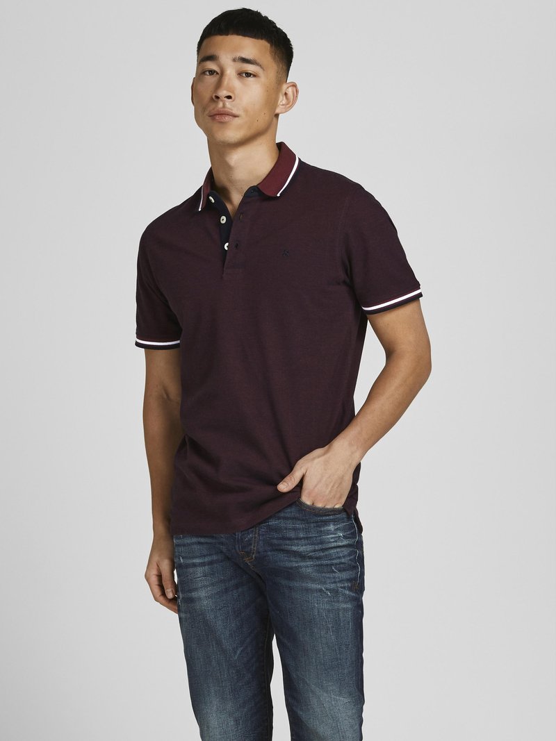 Burgundy-paitainen polo-paita, jossa on kontrastivärinen tummansininen kaulus ja valkoinen reunoitus, yhdistettynä sinisiin farkkuihin. Paitassa on kolmen napin suljin ja logo.