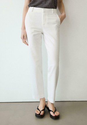 Osoba ubrana w białe, proste spodnie z wysokim stanem oraz czarne klapki na platformie, stoi z jedną ręką w kieszeni na jednolitym tle.