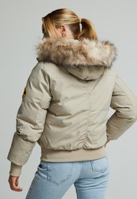 Béžová péřová bunda s kapucí lemovanou kožešinou. Disponuje přiléhavým střihem, žebrovanými manžetami a mírně nařaseným zadním švem. Měkká textura.
