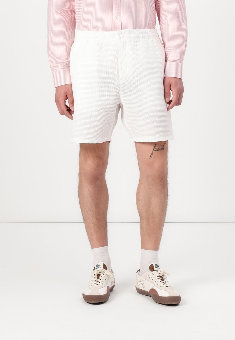 Pantaloni corti bianchi e texturizzati con vita elasticizzata, caratterizzati da un dettaglio con bottone e una vestibilità comoda, abbinati a scarpe e calze dai colori chiari.