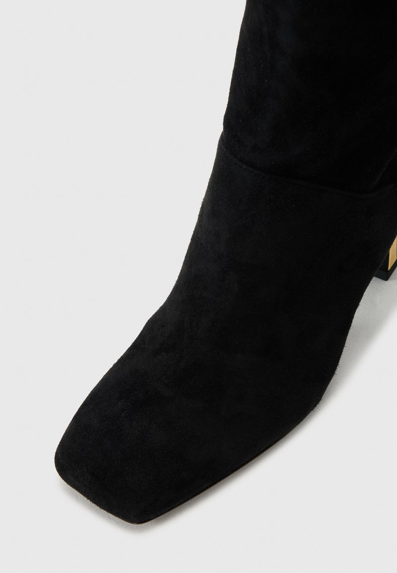 Schwarze Wildleder-Ankle-Boots mit einer quadratischen Zehenform und einem niedrigen Blockabsatz, der mit einem metallischen Akzent verziert ist. Die Textur wirkt weich und glatt.