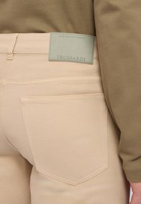 Pantalones vaqueros de mezclilla beige con un detalle en el bolsillo trasero y una etiqueta de la marca Trussardi de color gris colocada arriba. La parte superior es de algodón verde oliva.