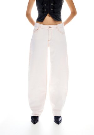 Ženska, oblečena v bele široke kavbojke z visokim pasom, črne čevlje z zoženim sprednjim delom in črno brezrokavnik, stoji z rokami za hrbtom.