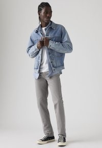 Νεαρός άνδρας με πλεξούδες, φορώντας γαλάζιο τζιν μπουφάν, λευκό πουκάμισο, γκρι παντελόνι και μαύρα αθλητικά παπούτσια, στέκεται μπροστά σε ένα απλό φόντο.