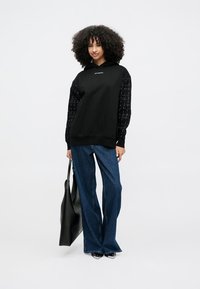 Μαύρο φούτερ με υφασμάτινα, διακοσμημένα μανίκια, συνδυασμένο με φαρδιά μπλε τζιν. Το μοντέλο κρατάει μια μαύρη τσάντα tote. Απλός και μοντέρνος σχεδιασμός.