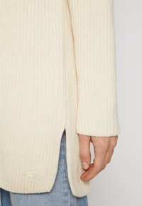 Nahaufnahme einer Hand neben dem seitlichen Schlitz eines cremefarbenen gestrickten Pullovers, der mit blauen Jeans getragen wird.