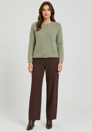 Femme portant un cardigan boutonné vert clair, un pantalon large marron foncé et des bottines marron foncé, debout devant un fond uni.