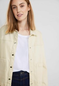Vaalea kerma-button-up takki, jossa on metalliset nepparit, kaksi rintataskua, käytetty valkoisen T-paidan ja tummansinisien farkkujen päällä. Sujuva pinta.