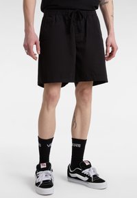 Pantaloni corti neri con vita elastica e cordoncino. Presentano tasche laterali e una vestibilità ampia. Indossati con scarpe da ginnastica nere e bianche e calze.