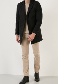 Cappotto nero in misto lana con revers a lancia, taschino sul petto e chiusura con bottoni, abbinato a pantaloni beige e stivaletti neri alla caviglia.