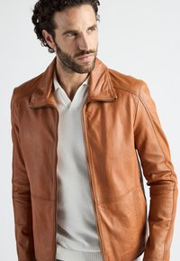 Brun læderjakke med lynlås, krave og sidelommer. Texturen er glat med sømdetaljer, parret med en hvid skjorte nedenunder.