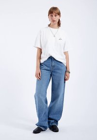 Weißes Baumwoll-T-Shirt mit einem Logo auf der Vorderseite, kombiniert mit locker sitzenden, hellblauen Jeans. Dazu schwarze Schuhe und eine silberne Halskette.