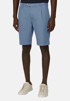 Mand iført lyseblå skræddersyede shorts med forlommer og beige sneakers, stående mod en ensfarvet baggrund.