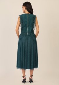 Türkises Spitzekleid mit gewellten Ärmeln und einem Reißverschluss hinten. Der Rock hat einen Pünktchen-Tüll-Überzug und endet auf halber Wade. Schwarze Absatzsandalen.