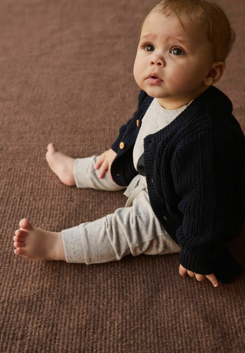 Cardigan en tricot bleu marine avec des boutons en bois, porté par-dessus un haut à manches longues gris clair et un pantalon gris à motifs. Bébé assis sur un tapis marron texturé.