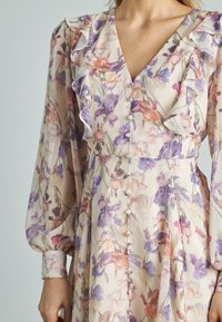 Vestido floral con escote en forma de V, volantes en la parte delantera y mangas largas abullonadas. Presenta flores en tonos pastel de color púrpura y melocotón sobre un fondo claro.