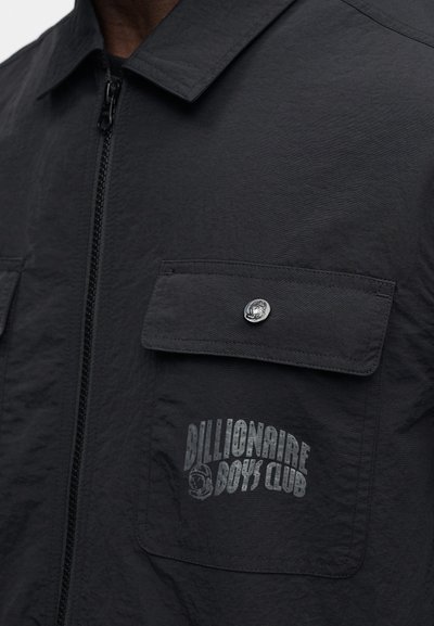 Μαύρο μπουφάν με φερμουάρ μπροστά, κατασκευασμένο από υφή υφάσματος, με δύο μπροστινές τσέπες με κουμπιά και τυπωμένο λογότυπο στην αριστερή τσέπη.