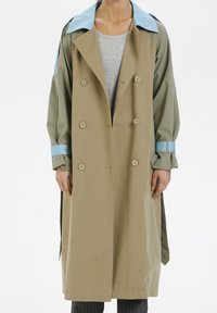 Lång beige trenchcoat med ljusblå krage och ärmsnoddar bärs över en grå ribbad tröja och mörkt randiga byxor.