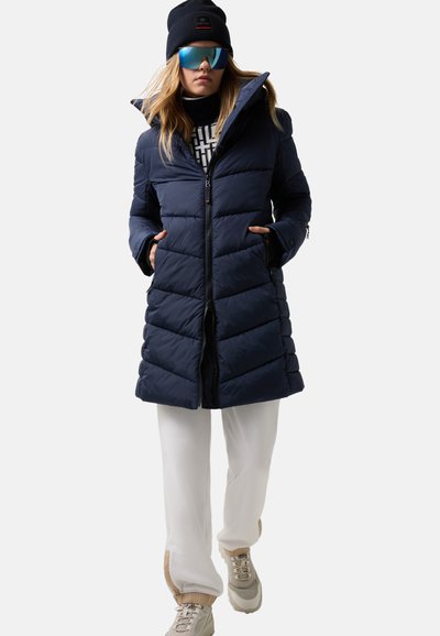 Mørkeblå puffjakke med høj krave, lynlås lukning og vandret syninger. Stylet med hvide bukser og beige sneakers. Inkluderer en hue og solbriller.