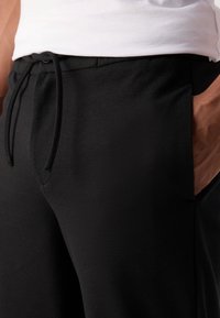 Svart joggebukser laget av glatt stoff, med elastisk midje og snor, samt sidelommer. Enkel og funksjonell design.