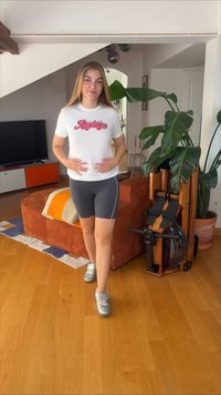 Νεαρή γυναίκα με αθλητική εμφάνιση περπατά μέσα σε εσωτερικό χώρο κοντά σε πορτοκαλί καναπέ, μεγάλο πράσινο φυτό και μηχάνημα κωπηλασίας με νερό σε σαλόνι.