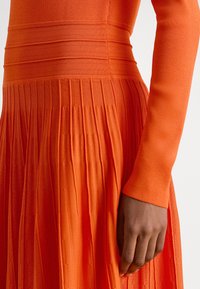 Dešinė ranka ilsisi šalia raukšlėto oranžinio megzto suknelės su horizontaliu rištu raštu viršutinėje kūno dalyje.