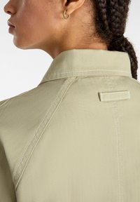 Chaqueta beige de espiga con cuello, que presenta costuras visibles y un detalle de lazo horizontal en la parte posterior.