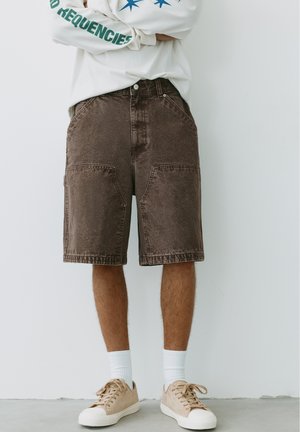 Personne portant un short en jean marron jusqu'aux genoux, des baskets beiges aux semelles blanches et des chaussettes blanches, debout contre un mur blanc uni.