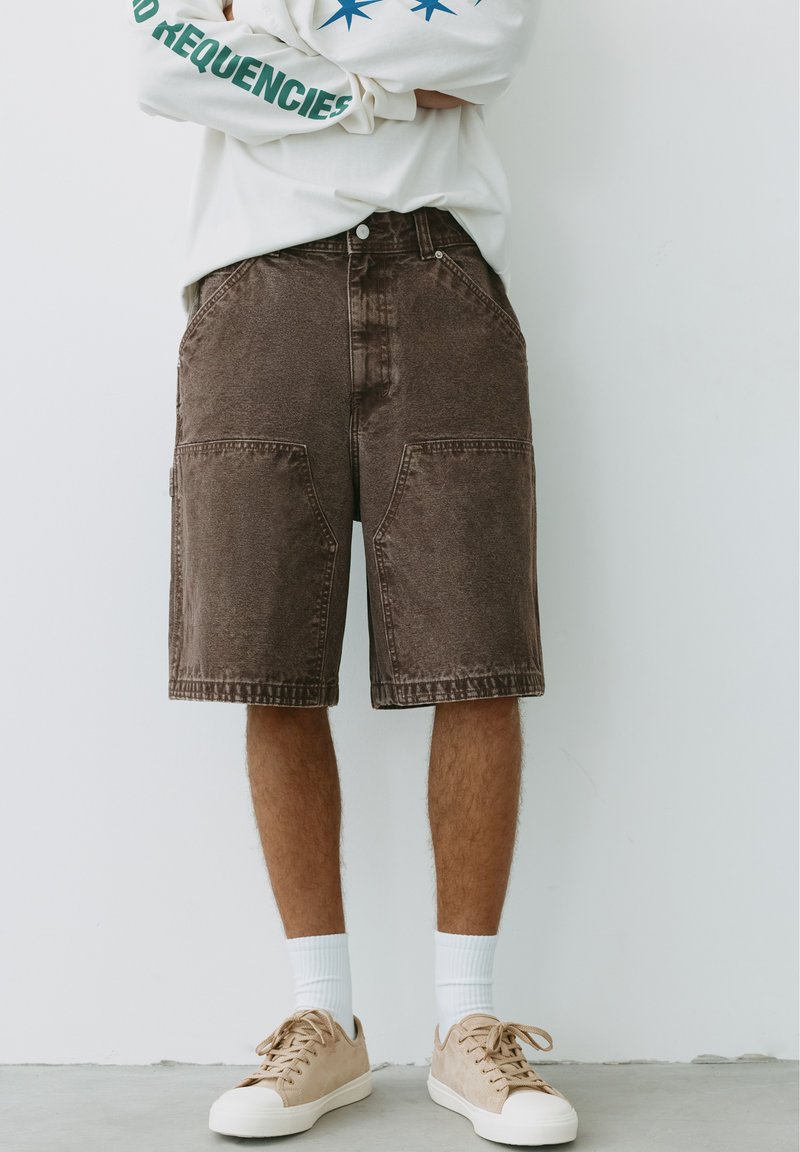 Personne portant un short en jean marron jusqu'aux genoux, des baskets beiges aux semelles blanches et des chaussettes blanches, debout contre un mur blanc uni.