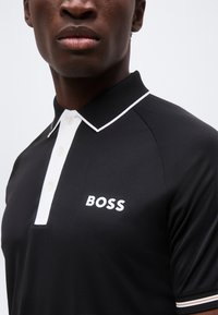Κοντινό πλάνο ενός ατόμου που φοράει μαύρο πόλο με λευκή επένδυση και το λογότυπο "BOSS" στο στήθος.
