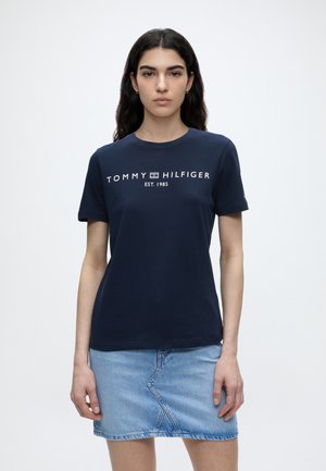 Kobieta z długimi ciemnymi włosami, ubrana w granatowy t-shirt Tommy Hilfiger i jasnoniebieską dżinsową spódnicę, stojąca na jednolitym białym tle.