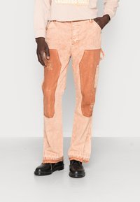 Persona que lleva pantalones de un naranja claro con parches de naranja más oscuro en los muslos, combinados con zapatos negros y una sudadera beige con texto.
